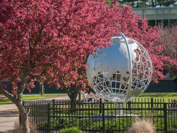 A photo of the silver, campus globe. Trees bud beside it, sprouting pink flowers. Green grass is behind the globe, and a black wrought-iron fence surrounds it.
