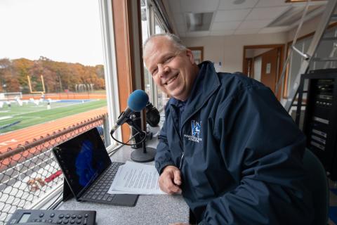 Man sits at a laptop with a microphone and notes in a booth overlooking the Westfield State University football field