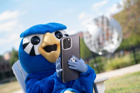 Nestor, the school mascot, wears his blue Owl suit. Nestor is holding a large phone in his hands and sits in front of the campus globe.