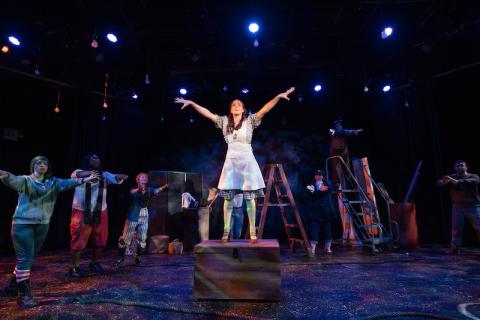 Westfield State's production of "Peter and the Starcatcher". A lone student wearing a blue dress is standing on top of a wooden chest on stage. Their hands and arms are splayed out above their head, at their sides. Several other actors surround them as they perform.