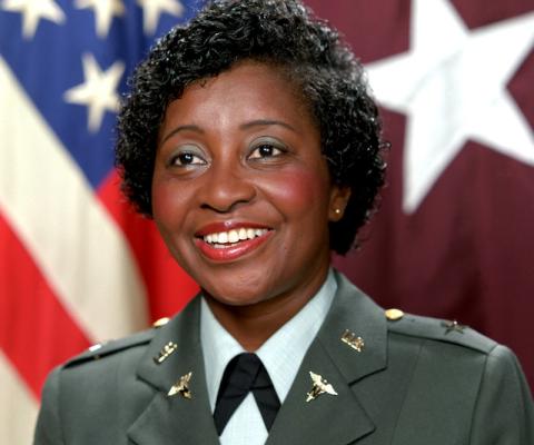 General Clara Adams-Ender, a United States General. She is an African American woman in an olive-green nurse uniform. She is wearing red lipstick and green eyeshadow, and standing in front of the American flag. Several gold pins decorate her uniform's lapel.