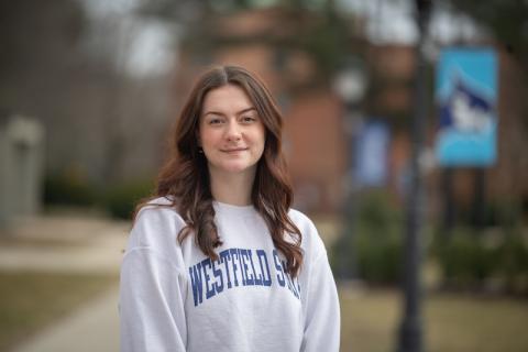 Paige Freeman, a senior at the University. She is outside, near the Ely building. She has long, brown hair and a gray sweatshirt which says "Westfield State" in blue lettering. Blurred out trees, grass, and the sidewalk are in the background.