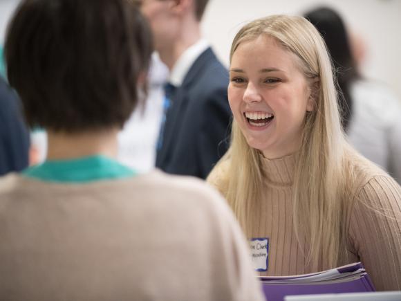 Female student in tan turtleneck sweater, smiles laughs with a prospective employer at the career fair