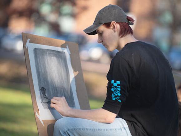 A student uses their hand to blend a charcoal drawing while seated outside in a class setting.