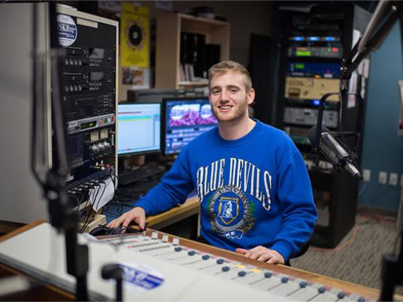 A student smiles while seated at a soundboard in a studio setting.