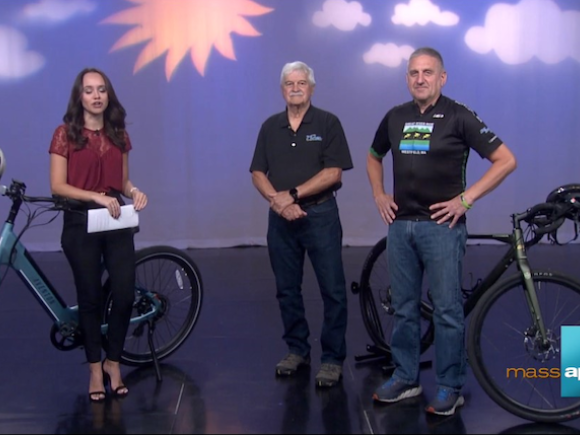 Kaylay Hevey, a newsanchor with long, brown hair, a red shirt, and black pants, stands in front of a bicycle and purple backdrop as she stands next toDon Podolski, Director of Education, and Paul Cacolice, a board member for the Great River Ride. 