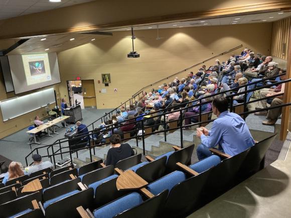 A photo of the Savignano Auditorium in Wilson Hall, with people sitting in the blue-cushioned seats. They watch a panel of guest-speakers discuss clean energy in front of them.