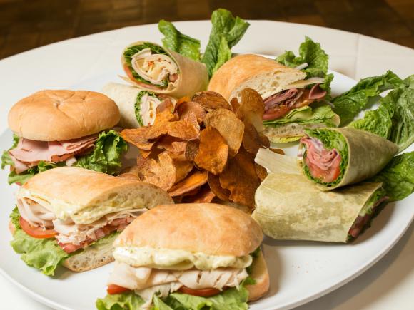 Sandwich platter featuring turkey ciabatta sandwiches and wraps with home made chips in the center on white plate.