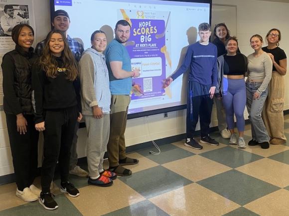 Westfield State University class of Advanced Public Relations pose in front of a poster design for “Stick Together with Rick’s Place Where Hope Scores Big,” campaign. 