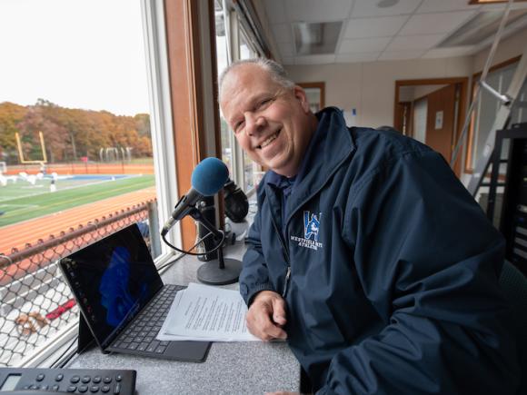 Man sits at a laptop with a microphone and notes in a booth overlooking the Westfield State University football field