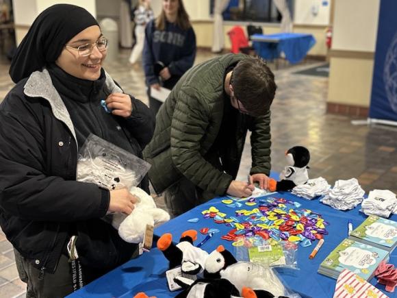 Two students stand in front of a table decorated with black and white penguin plushies, stickers, white socks, and heart-shaped pins. This is for Winter Weekend on campus, where students could make their own winter survival kits for the cold weather.