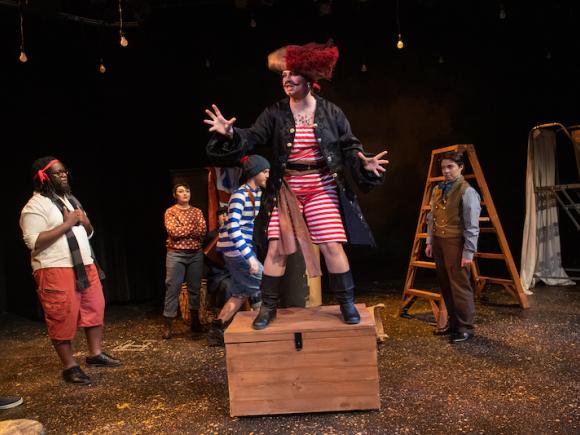 Westfield State's production of Peter and the Starcatcher. An actor is standing on a wooden treasure chest atop a stage, wearing a pirate's costume. Several other actors in costumes surround them.
