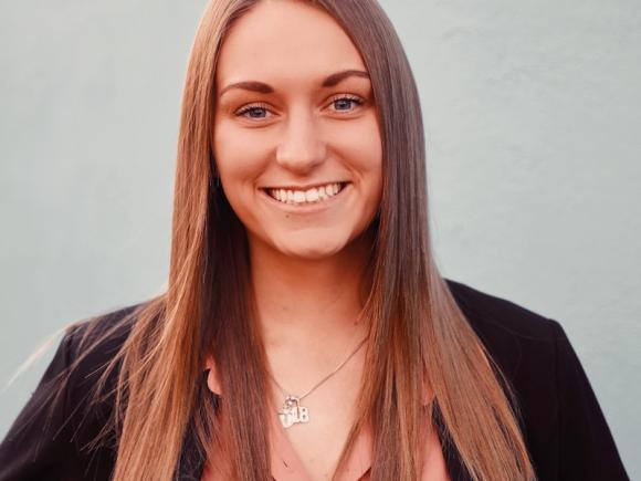 Rachel Gelina, class of 2020. She is a long and brown-haired white woman and wears a black jacket with a peach undershirt. She is standing in front of a gray, nondescript background and smiling at the camera.