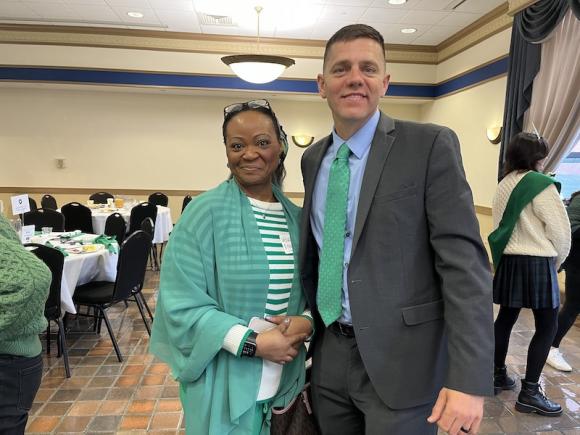 Senator John Velis and an attendee of this year's Chamber of Commerce breakfast. Her name tag is unclear. He is dressed in a suit with a green tie, and she wears a white-and-green sweater along with a sheer, green shawl. Black glasses sit atop her head. In the background, white-clothed tables full of plates and food rests and one of the Colleen's Court women from Holyoke stands off to the side as she talks to someone else.