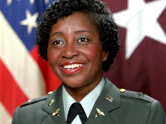 General Clara Adams-Ender, a United States General. She is an African American woman in an olive-green nurse uniform. She is wearing red lipstick and green eyeshadow, and standing in front of the American flag. Several gold pins decorate her uniform's lapel.