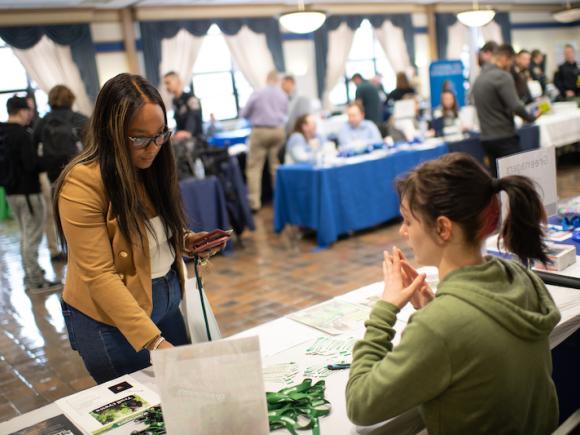 The career fair, 2024. The photo is of a white-clothed booth with green pamphlets and brochures on it. A young woman in an olive green sweatshirt and a ponytail sits behind it. In front, a young African American student in a mustard yellow, long-sleeve jacket and jeans touches one of the brochures. She also wears black glasses. In the background, there's blurred out people, booths, and spectators enjoying the booths set up for the career fair.