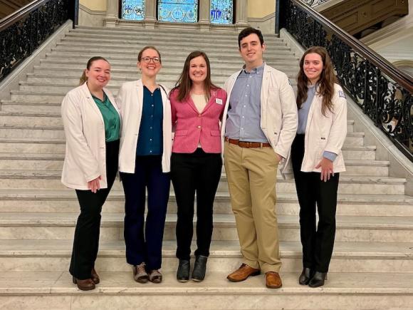 Victoria Farrara-Lawlor, Edward Krajkowski, Caitlyn Grahn, and Alexis Bergman, and Susan McDiarmid at the 2024 PA Lobby Day Conference. They pose and gather on a large staircase, and wear white coats. Susan, however, wears a pink coat.