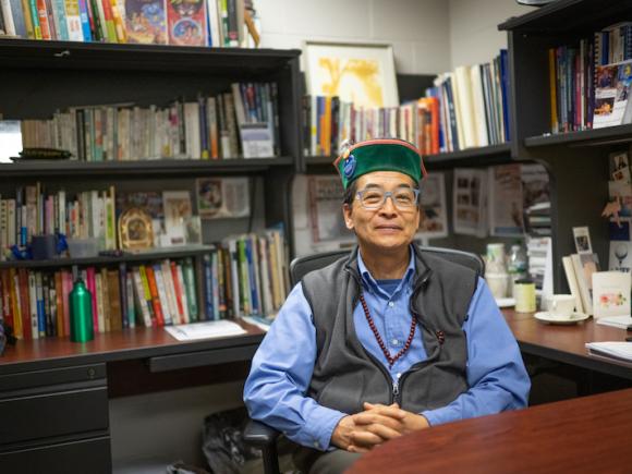Professor Max Saito of the Communication Department. He sits in his office, which is walled with several shelves of books. His hands are clasped over his torso, and he smiles at the camera in a vest and green hat.