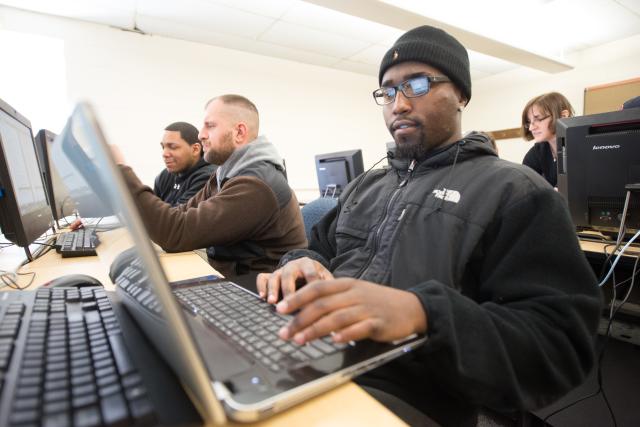 Male student in glasses and beanie cap working on laptop in class