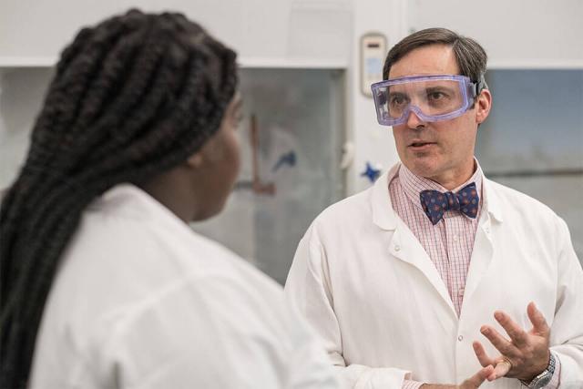 A professor wearing a lab coat and safety goggles talks with a student in a laboratory setting.