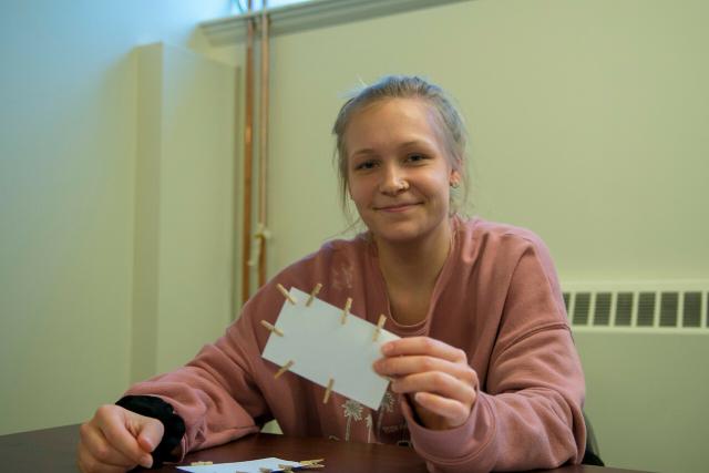 Health Sciences student wearing pink shirt smiling holding index card with clothes pin on it for interactive classroom project.