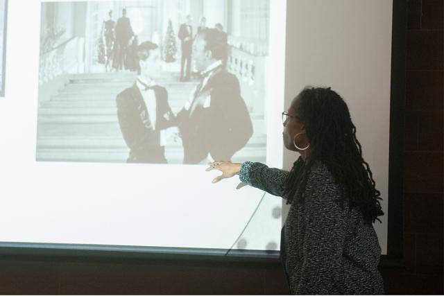 An ethnic and gender studies professor motions at an image from a black and white movie being projected on a screen.