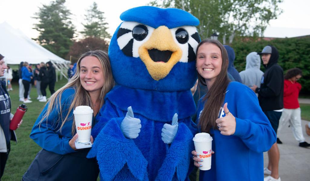 Two students with Nestor mascot at pep rally wearing blue shirts holding Dunkin cups with thumbs up.
