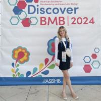 Taylor Camossi at the 2024 BMB Biochemistry Conference in San Antonio. She poses in front of a large white, pink, and blue poster which says, "Discover BMB 2024". She is wearing a white and black dress, white shoes, and a blue lanyard. Graphics of flowers and cells dot the poster behind her.