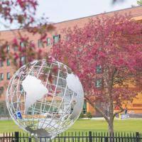 A stock photo of the campus globe. Two pink-flowered trees rest beside the globe, and University Hall can be seen in the background.