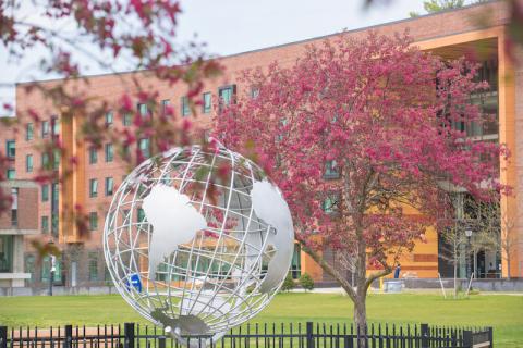A stock photo of the campus globe. Two pink-flowered trees rest beside the globe, and University Hall can be seen in the background.