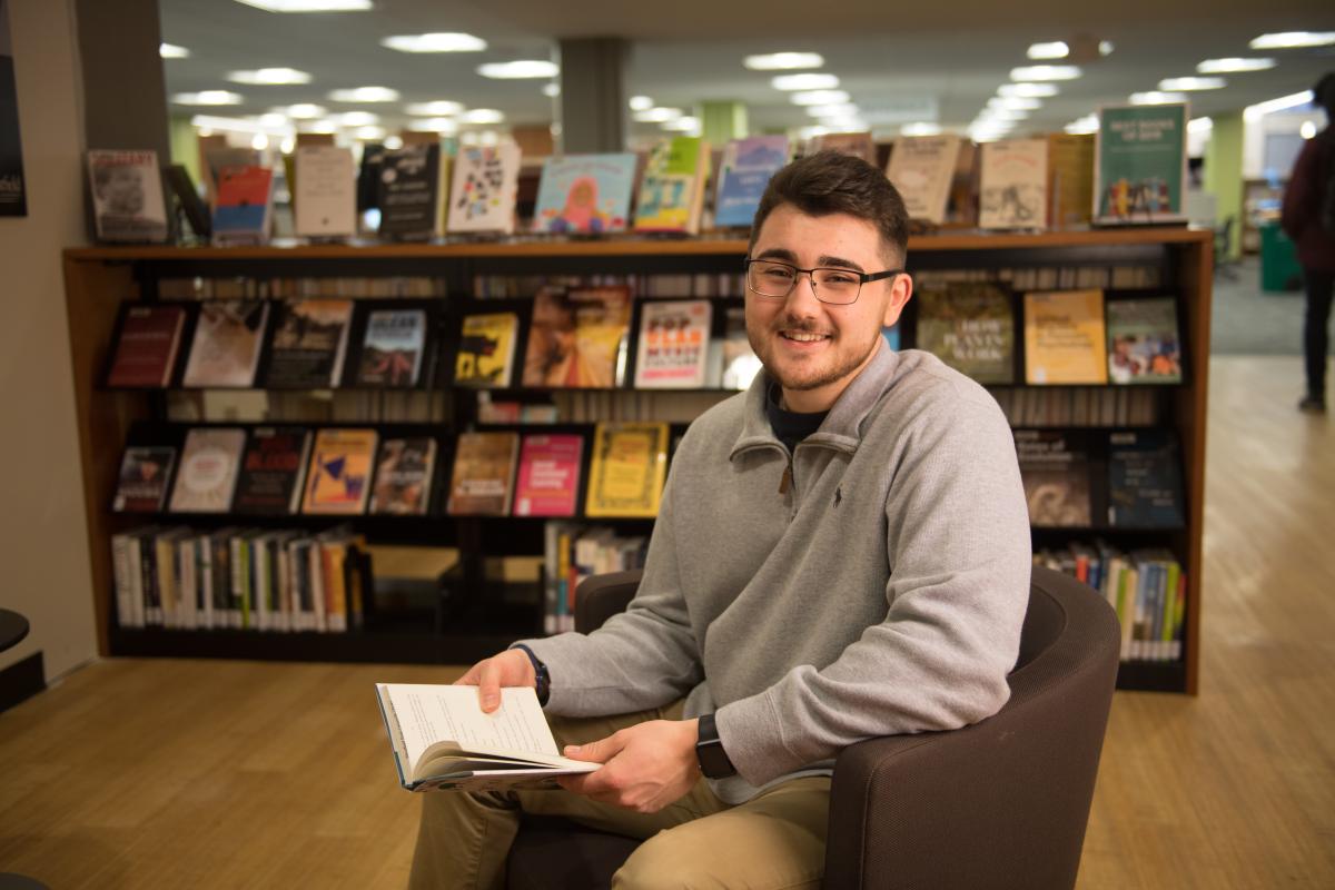 make student in glasses sitting in the Ely Library with a book in his lap