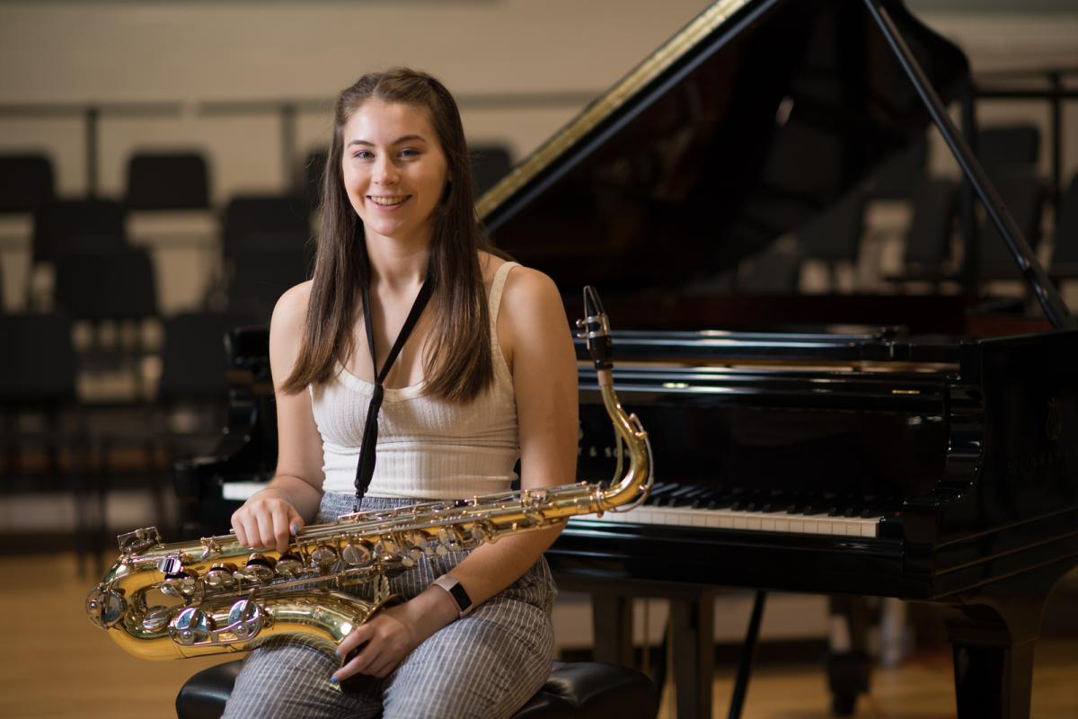 Music student sitting on a piano bench with a piano behind her holding a saxophone.