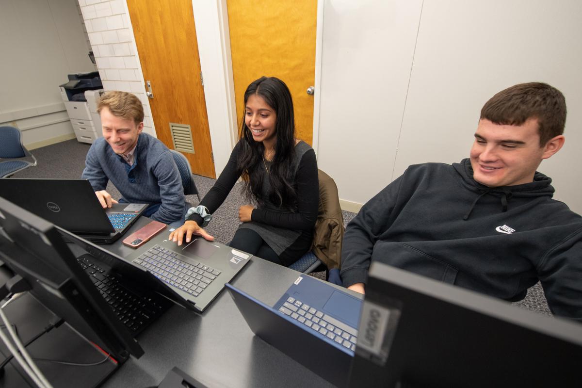 Three data science students in classroom in front of desk top computers smiling