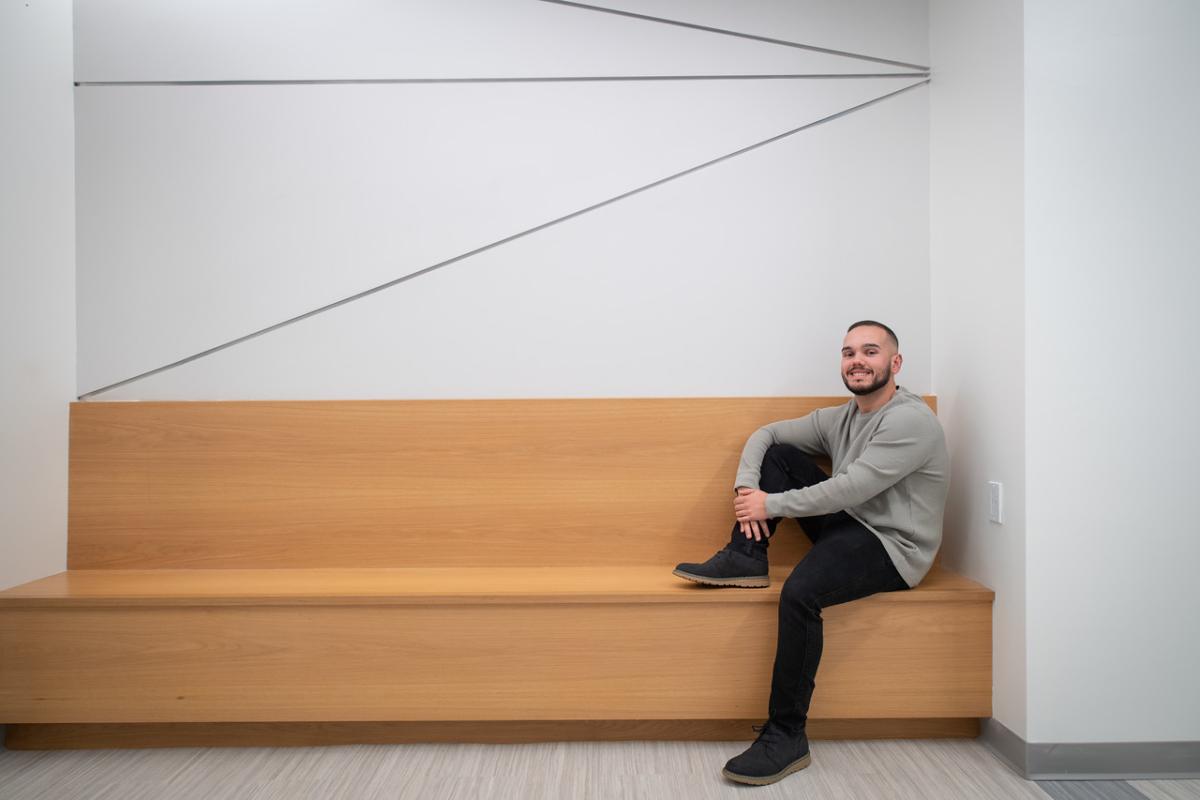 Nate a student in the reading specialist program sitting on a bench with a white wall with black lines behind him.