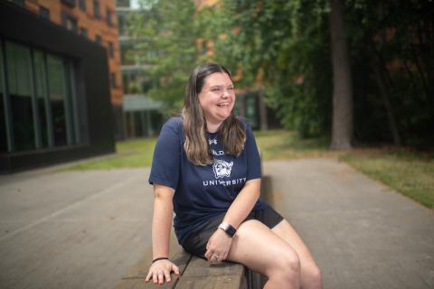 Emily Czupta, a brown-haired, white woman wearing a navy blue T-shirt and black shorts smiles to the side. She sits on a stone bench near a brick building. Green trees dot the background, which is blurred.