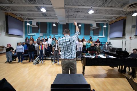 A chorus of various WSU students is facing a professor in a blue-plaid button down as he lifts his arms in the air and conducts them.