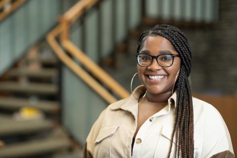 Sydney Maiden, a young, African American woman dressed in a beige, button-down short sleeve shirt. She also wears black glasses and hoop earrings. Her hair is swept into a side ponytail.