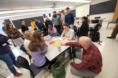 Students gather around a white table with pink and yellow pieces of paper on it. Dr. Moore, in a red and black shirt, crouches next to them.
