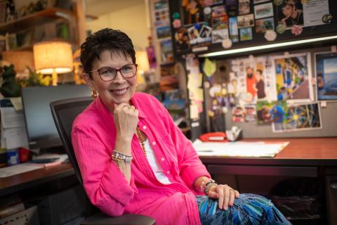 Regina, wearing a pink jacket, bracelets, earrings, glasses, and blue pants, is sitting in her office with her hand tucked under her chin and smiling.
