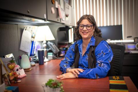 Estelle Camacho of Student Accounts. She is dressed in a long-sleeve blue shirt, blue glasses, and sits at her desk.