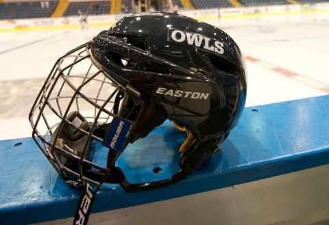 A close-up shot of a navy blue hockey helmet that has "Owls" printed in white letters on its side. It's resting on the edge of the wall of a hockey rink.