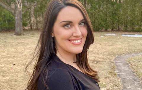 Desiree Nickel '11, a woman with long, brown hair, smiles with pine trees from an unnamed forest in the background.