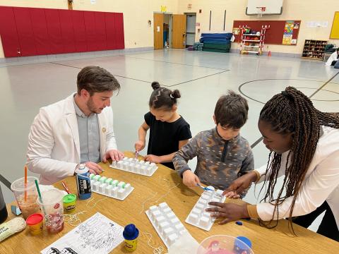 Two students in white lab coats present to the kids at Paper Elementary School in Westfield. The students and two children sit at a wooden desk and play with ice-cube trays as part of a dental hygiene demonstration.