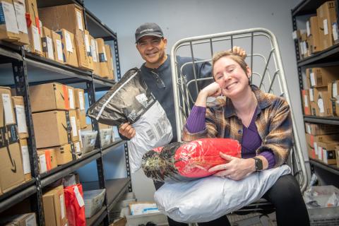 Mike DeJesus and Natalie Lussier of Mail Services, sitting on a package cart and smiling at the camera. They are surrounded by stacks of packages around them, displaying the amount of parcels needing distribution across campus.