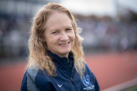 Nancy Bals, Associate Director of Athletics, smiles and poses on the University's track, which is blurred out. She is wearing a long-sleeved, navy sweater with the University's Owl logo on it.
