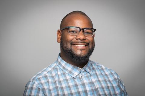 Joseph Seal, Academic Support Coordinator for the Course Achievement, Retention, and Engagement Center. He wears a white and blue-checkered button-down shirt and black glasses. He smiles in front of a plain, gray background.