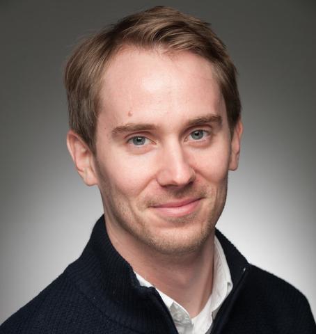 Colin Moge, class of 2016. He smiles in front of a plain, gray background, as part of his headshot. He is wearing a black sweater with a white shirt underneath. He has short, blond hair and blue eyes.