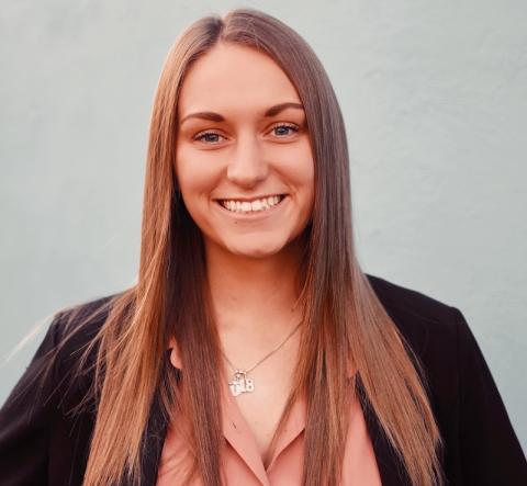 Rachel Gelina, class of 2020. She is a long and brown-haired white woman and wears a black jacket with a peach undershirt. She is standing in front of a gray, nondescript background and smiling at the camera.