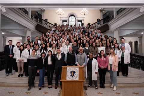 2024 Lobby Day, an event for Physician Assistants and students studying to be PAs. A large group of people, some in white coats, gather on the steps of the venue in Boston, MA. A wooden podium sits before all of them. Two chandeliers hang in the background behind them.