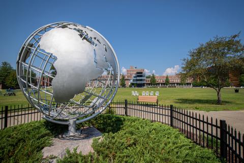 A stock photo of the silver campus globe. The sky above is blue, with green grass and vegetation around he globe itself, wrapped in by a black fence. Ely is in the distance, with a tree to the side of the frame.